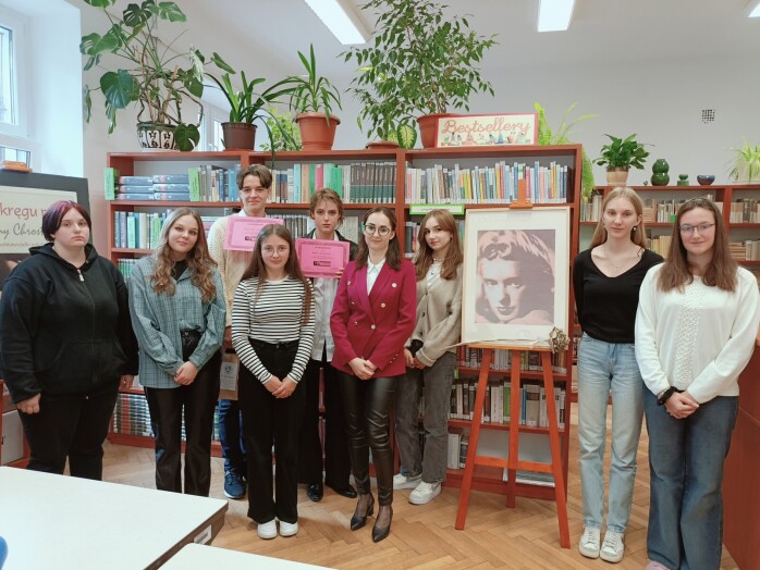 Grupa uczestników konkursu. W tle regały z książkami. Obok portret Grażyny Chrostowskiej.