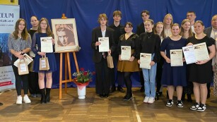 21.	Grupa młodych uczestników konkursu wraz z członkami jury, w środkowej części portret i bukiet czerwonych róż.