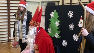 Na krześle siedzi św. Mikołaj, towarzyszą mu dwie uczennice IV Liceum. Dziewczyny mają na głowach czerwone czapki. Jedna z nich w ręku trzema prezent, a druga pastorał św. Mikołaja.