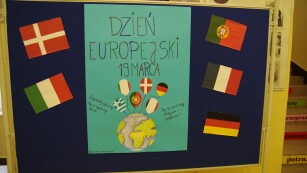 Granatowa tablica stoi na szkolnym korytarzu. Na niej zawieszony duży karton w kolorze niebieskim. Na dole widać kulę ziemską, do której przypięte są kolorowe balony – mają barwy flagi różnych państw europejskich. Powyżej napis - hasło wydarzenia wraz z datą. Po prawej stronie plakatu zawieszona jest flaga Portugalii, Francji i Niemiec. Po stronie lewej – flaga Danii i Włoch.