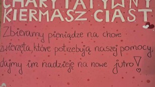Zdjęcie ręcznie wykonanego plakatu w kolorze różowym, białe litery w tekście charytatywny kiermasz ciast, czarne litery w tekście zachęcającym do ofiarności
