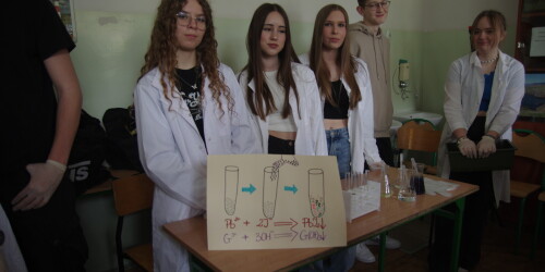 Trzy dziewczęta w białych fartuchach lekarskich, przed nimi stolik z odczynnikami i probówkami i młodzieniec w bluzie kremowej
