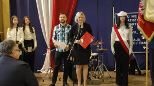 Dziewczyna wbiałej bluzce, mężczyzna w koszuli w kratę, kobieta w ciemnym kostiumie, dziewczyna reprezentująca sztandar szkoły