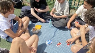 Grupa młodych ludzi siedzących na kocy, w rękach karty do gier planszowych